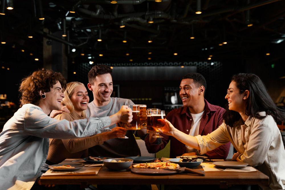A group of friends laughing and raising their glasses for a toast at a long wooden table during a Friendsgiving dinner at a bar.