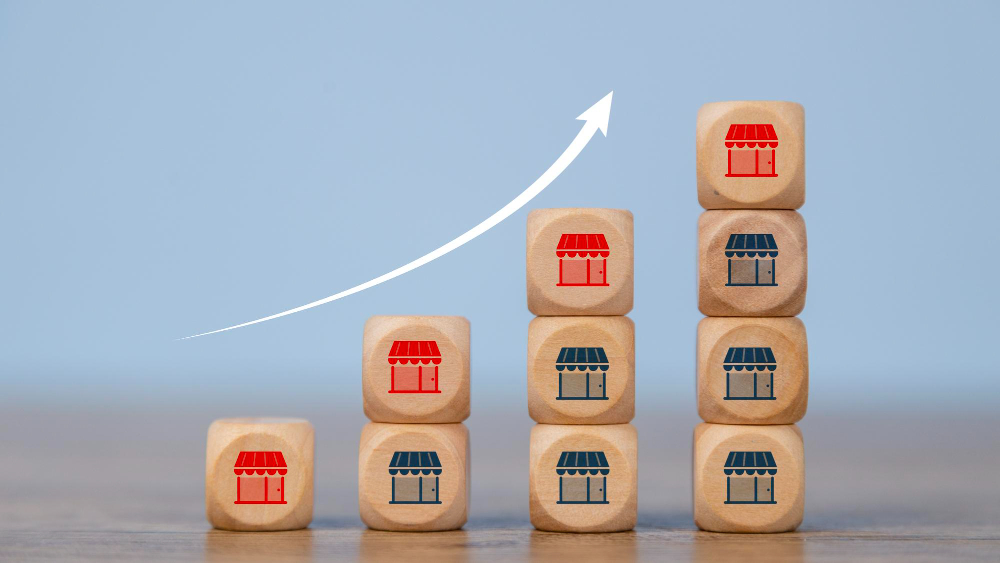 A bar graph made of wooden blocks with storefront icons, showing a white arrow pointing up, symbolizing business expansion.