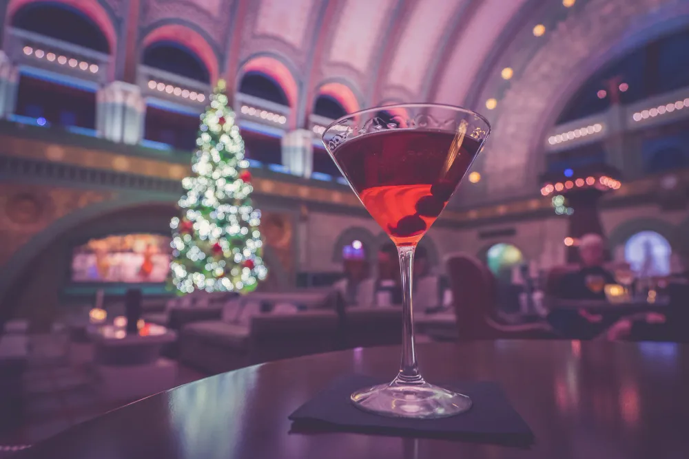 A red holiday cocktail in a martini glass on a dark wood table, with a festive Christmas tree blurred in the background of an elegant bar.