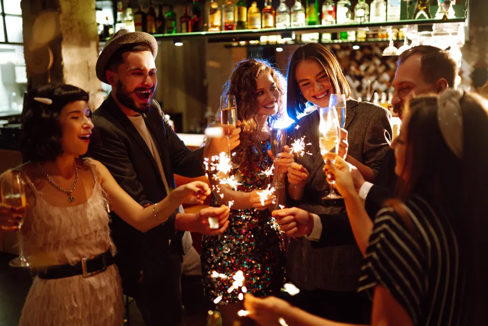 A group of five friends celebrating in a dimly lit bar, holding lit sparklers and champagne flutes, laughing and smiling.