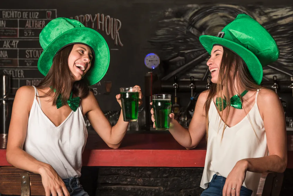 Two smiling women wearing green top hats and bow ties toast with glasses of green beer at a rustic Texas bar counter.
